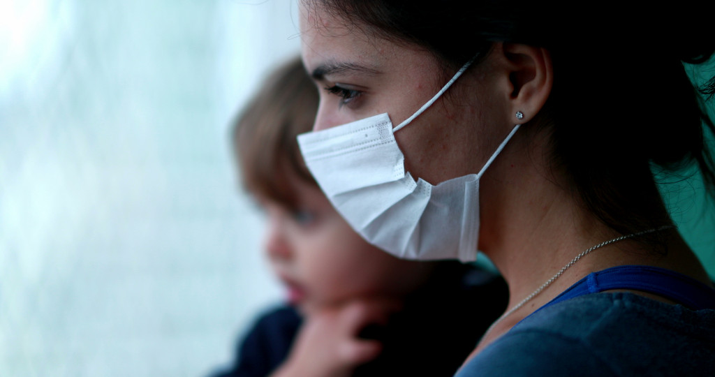 Mother wearing covid-19 mask holding baby looking outside standing by window