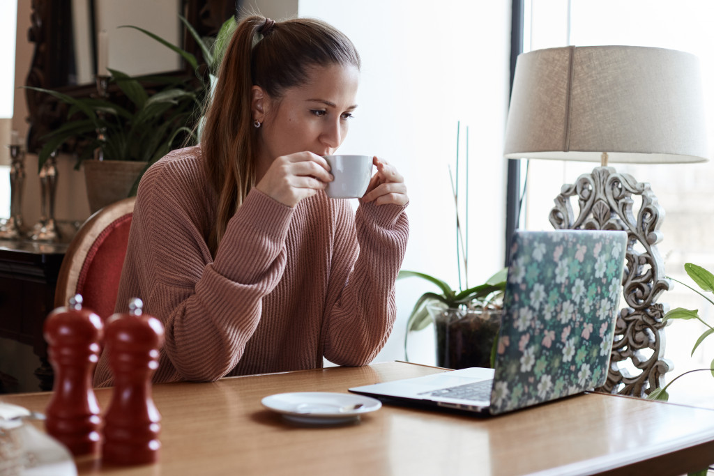 Young attractive Caucasian brown-eyed female in casual sweater is sitting at the restaurant, holding the cup of hot beverage and watching movie or video on her modern portable computer.