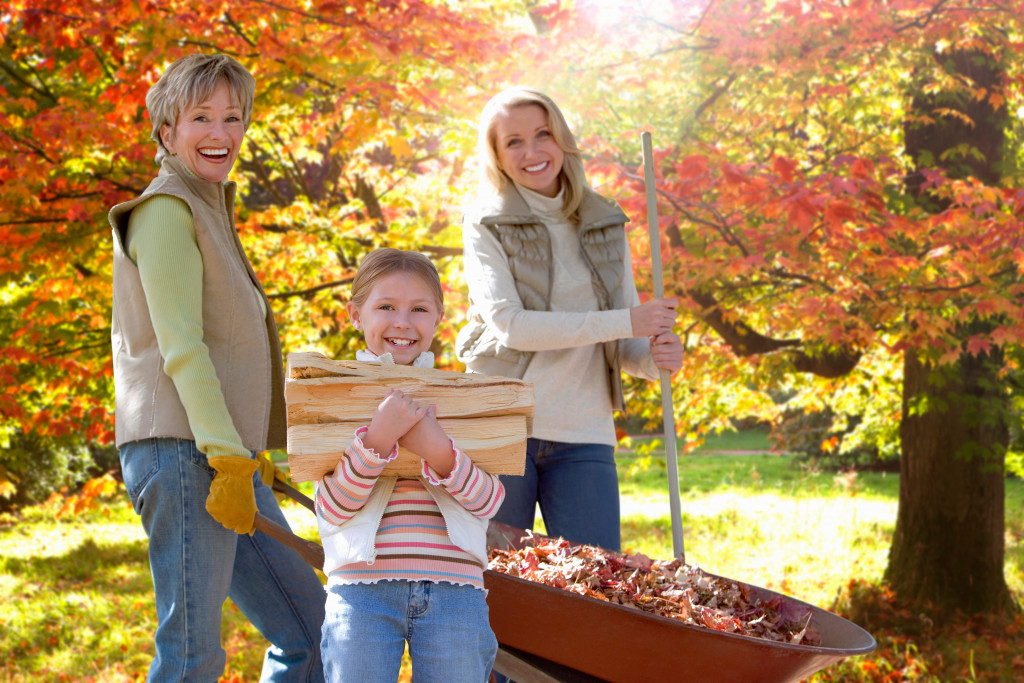 Whole family doing yard work. A young girl holding a log of woods in front while her mother and grandmother are collective leaves from the yard