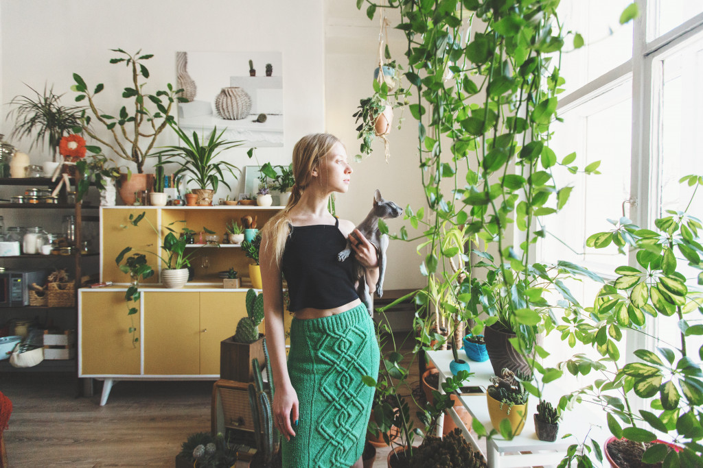 Young woman looking outside a window with her cat in a room filled with indoor plants.